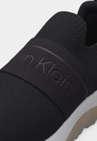 Baskets noires à enfiler avec tige en maille respirante, bande élastique sur le dessus embossée du logo "Calvin Klein", et semelle blanche et beige.