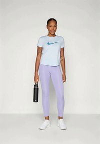 Šviesiai mėlynas trumpomis rankovėmis marškinėliai su turkio spalvos Nike logotipu, derinami su levandų spalvos leggingais. Laiko juodą vandens buteliuką. Baltos sportinės bateliai.
