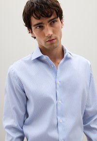 Un joven vestido con una camisa de botones blanca y azul claro, con rayas verticales, mirando ligeramente hacia un lado contra un fondo simple.