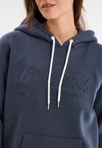 Haut à capuche bleu marine avec un logo texturé, cordons blancs et accents métalliques dorés. Il dispose d'une poche avant et d'un tissu doux et lisse.