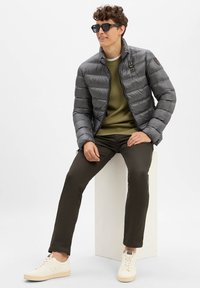 Grau gesteppte Pufferjacke mit horizontalen Nähten, grüner gerippter Pullover, dunkle Hosen und helle Sneakers. Zubehör umfasst schwarze Sonnenbrillen.