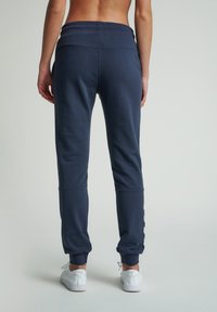 Tmavě modré tepláky s úzkým střihem, elastickým pasem a žebrovanými lemy. Mají bílý vzor loga na boku a hladký povrch.