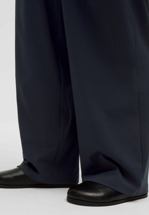 Široke mornarske hlače prekrivajo črne ravne čevlje, prikazano od spodnjega dela noge navzdol na enostavni ozadju.