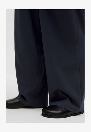 Široke mornarske hlače prekrivajo črne ravne čevlje, prikazano od spodnjega dela noge navzdol na enostavni ozadju.