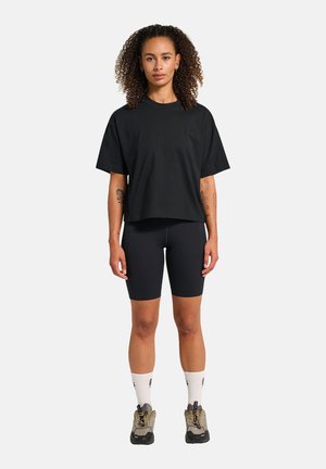 Frau mit lockigem Haar steht frontal und trägt ein lockeres schwarzes T-Shirt, schwarze Fahrradhosen, weiße Socken und beige Wanderschuhe.