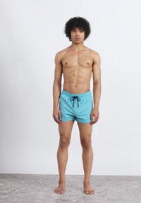 Vīrietis ar cirtainiem matiem stāv basām kājām, ģērbies gaiši zilās peldbiksēs ar melnu auklu, pret vienkāršu fona.