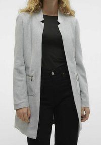 Světle šedý blazer v herringbone vzoru, s jedním řadovým zapínáním a dvěma předními kapsami se stříbrnými zipy, nošený přes černý top a černé kalhoty.