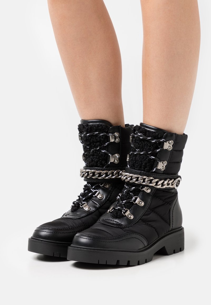 Bottines noires matelassées avec semelles épaisses, détail chaîne argentée autour des chevilles et lacets noirs et blancs aux pieds d'une personne.