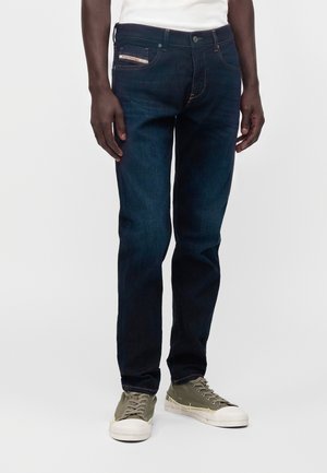 Homme portant un jean bleu foncé et des baskets vert olive debout devant un fond blanc.
