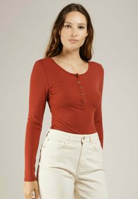 Bodysuit à manches longues en ribbed couleur rouille avec un col rond, détail de boutons sur le devant et une coupe ajustée, associé à un bas en denim de couleur claire.