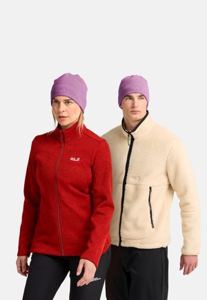 Frau in roter Fleecejacke und schwarzer Hose und Mann in beiger Fleecejacke und schwarzer Hose, beide tragen lila Mützen, stehen nebeneinander.