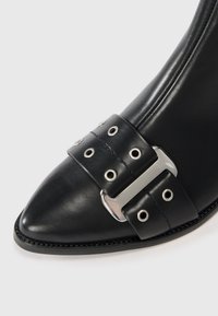 Bota de tornozelo em couro preto com biqueira pontiaguda, apresentada com uma faixa larga com acessórios de metal e ilhós circulares, finalizada com costura contrastante.