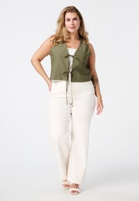 Olijfgroen cropped vest met een striksluiting, gecombineerd met hoge crèmekleurige wijde broeken en gevlochten haksandalen. Neutrale achtergrond.