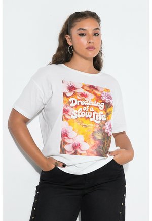 Jeune femme aux longs cheveux portant des boucles d'oreilles créoles, un t-shirt blanc à motif floral avec texte, et un pantalon noir avec des boutons dorés, mains dans les poches.