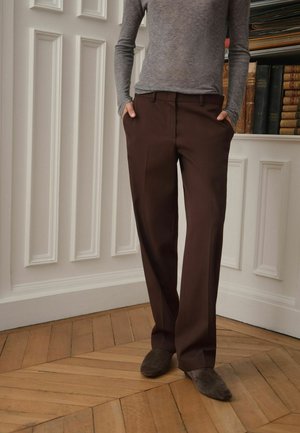Persona che indossa pantaloni marroni su misura, maglia grigia a manica lunga e scarpe marroni, in piedi su pavimento in parquet vicino a una parete bianca con pannelli con libri.
