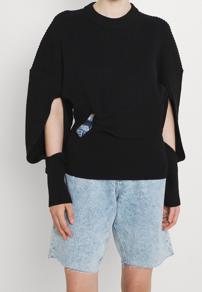 Black ribbed sweater with wide sleeves and a knotted detail at the waist, paired with light denim shorts.