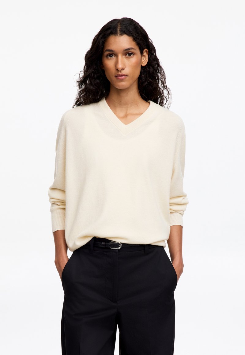 Femme aux longs cheveux bouclés portant un pull crème à col en V et un pantalon noir, les mains dans les poches, sur un fond blanc uni.