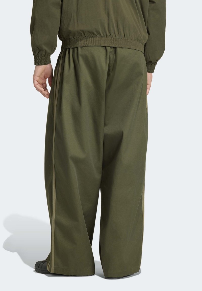 Personne portant un pantalon large vert olive et une veste assortie, debout avec les mains détendues le long du corps sur un fond uni.