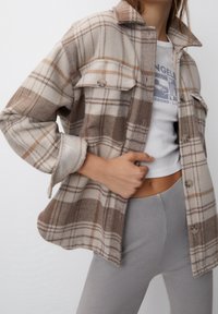 Brązowo-beżowa koszula typu shacket w kratę z dużymi patkowymi kieszeniami, zapięciem na guziki i miękką fakturą. Noszona na białym t-shircie z nadrukiem.