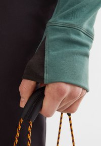 Langarmoberteil mit einem tiefblauen Ärmel und dunklem Akzent; strukturierter Stoff; Hand, die einen schwarzen Griff mit mehrfarbigem Kordel hält.