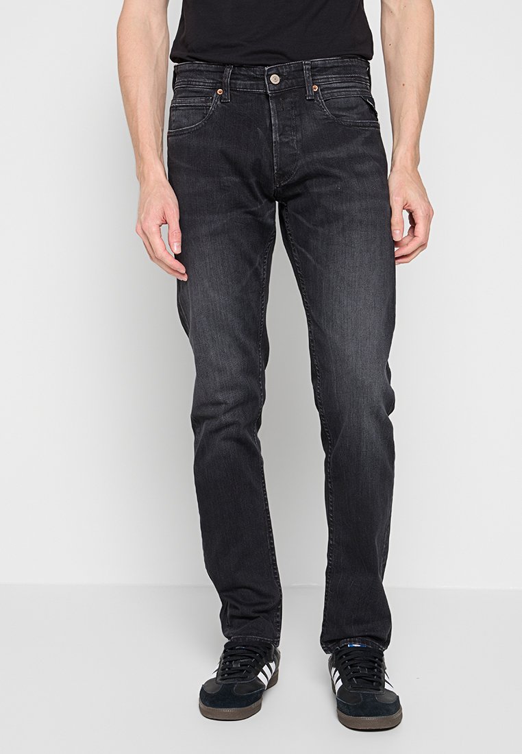 Man in slim-fit zwarte jeans en zwarte sneakers met witte strepen, staand tegen een witte achtergrond.