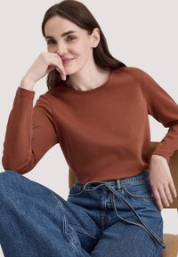 Bruine lange mouwen shirt met een ronde halslijn, gecombineerd met wijde lichtblauwe spijkerbroek met een trekkoord, zittend op een licht houten stoel.