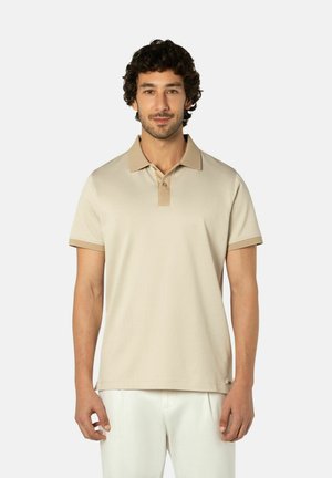 Man met krullend donker haar draagt een beige poloshirt met een donkerdere kraag en mouwen, gecombineerd met een witte broek, staand tegen een effen achtergrond.