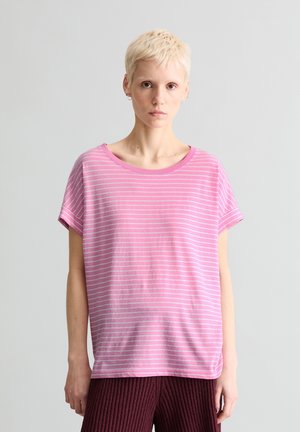 Personne aux cheveux blonds courts portant un t-shirt rayé rose et blanc et un pantalon texturé foncé, debout devant un fond clair uni.