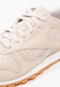 Gros plan sur une basket beige Reebok avec une tige texturée, des lacets en tissu et une semelle en caoutchouc marron clair sur fond blanc.