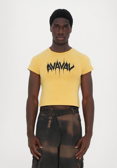 Gul cropped t-shirt med sort grafisk tekst "AVAVAL." Korte ærmer og et tætsiddende design. Båret sammen med sorte slidte cargo-bukser.