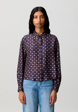 Femme aux longs cheveux noirs portant une chemise à motifs bleu foncé boutonnée et un jean bleu clair, debout devant un fond gris uni.