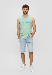 Hellgrünes ärmelloses Shirt mit blauer Grafik, kombiniert mit hellblauen Denim-Shorts und weißen Sneakers mit schwarzen Akzenten.