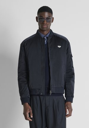 Hombre con chaqueta bomber oscura, camisa azul marino, corbata a rayas, pantalones de rayas finas y gafas de sol oscuras, de pie frente a un fondo gris liso.