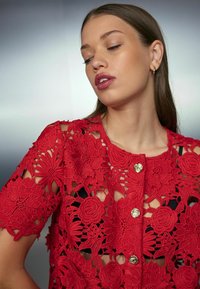 Sarkana mežģīņu blūze ar ziedu izšuvumiem, zelta pogām, īsām piedurknēm un izgriezumiem, veidojot teksturētu, atklātu dizainu.