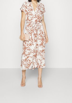 Kvinde iført en brun og hvid mønstret midikjole med bindebånd i taljen, holder en beige clutch og har gennemsigtige remsko på.