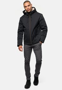 Chaqueta con capucha negra, cierre frontal de cremallera, forro texturizado y dos bolsillos frontales; combinada con pantalones gris ajustados y calzado negro.