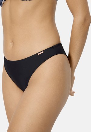 Mujer con bikini negro con una pequeña etiqueta metálica de marca en la cadera izquierda, sobre un fondo liso y claro.