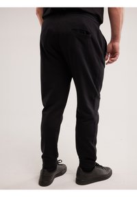 Mees, kes kannab musti kitsaid vabaajapükse ja musti tossusid, seisab heledal taustal, käed lõdvestunult kõrval.