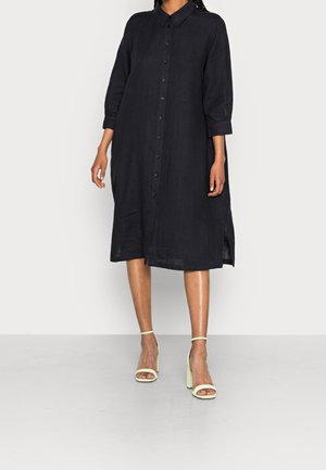 Femme portant une robe midi noire ample à boutons avec des manches trois-quarts et des sandales à talons hauts jaune pâle sur un fond uni.