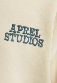 Beige fabric with raised blue embroidery reading "APREL STUDIOS", showcasing a textured surface and clean, modern font design.