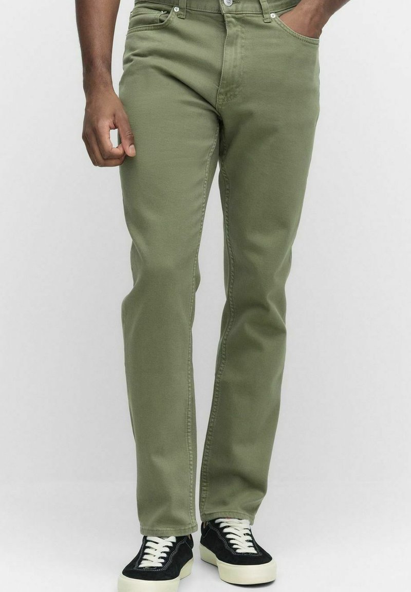Olivgröna jeans tillverkade av bomull, med rak skärning och en slät textur. Modellen bär svarta sneakers med vita sulor.