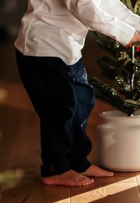 Tummansiniset housut, joissa on rento malli ja kerätyt lahkeet, yhdistetty valkoiseen nappipaitaan. Paljain jaloin puulattialla pottipuun vieressä.