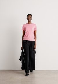 Pink bomulds t-shirt, korte ærmer, rund halsudskæring, båret med løse sorte bukser. Model holder en sort taske og står foran en neutral baggrund.
