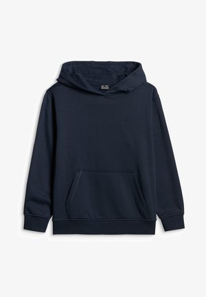 Garçon debout les mains dans les poches, portant un sweat à capuche bleu foncé, un pantalon cargo gris et des baskets blanches sur un fond gris uni.