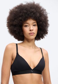 Νεαρή γυναίκα με φουντωτά σγουρά μαλλιά, φορώντας μαύρο αθλητικό σουτιέν με το λογότυπο Puma, σε απλό λευκό φόντο.