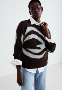 Brun og grå mønstrete genser med en myk, strukturert overflate, som har et karakteristisk virvelmønster. Båret over en hvit skjorte og kombinert med lyse jeans.