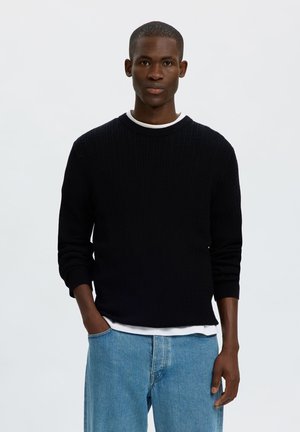 Ung mand står med hænderne i lommerne iført en sort sweater, hvid undertrøje og lyseblå jeans mod en ensfarvet lys baggrund.