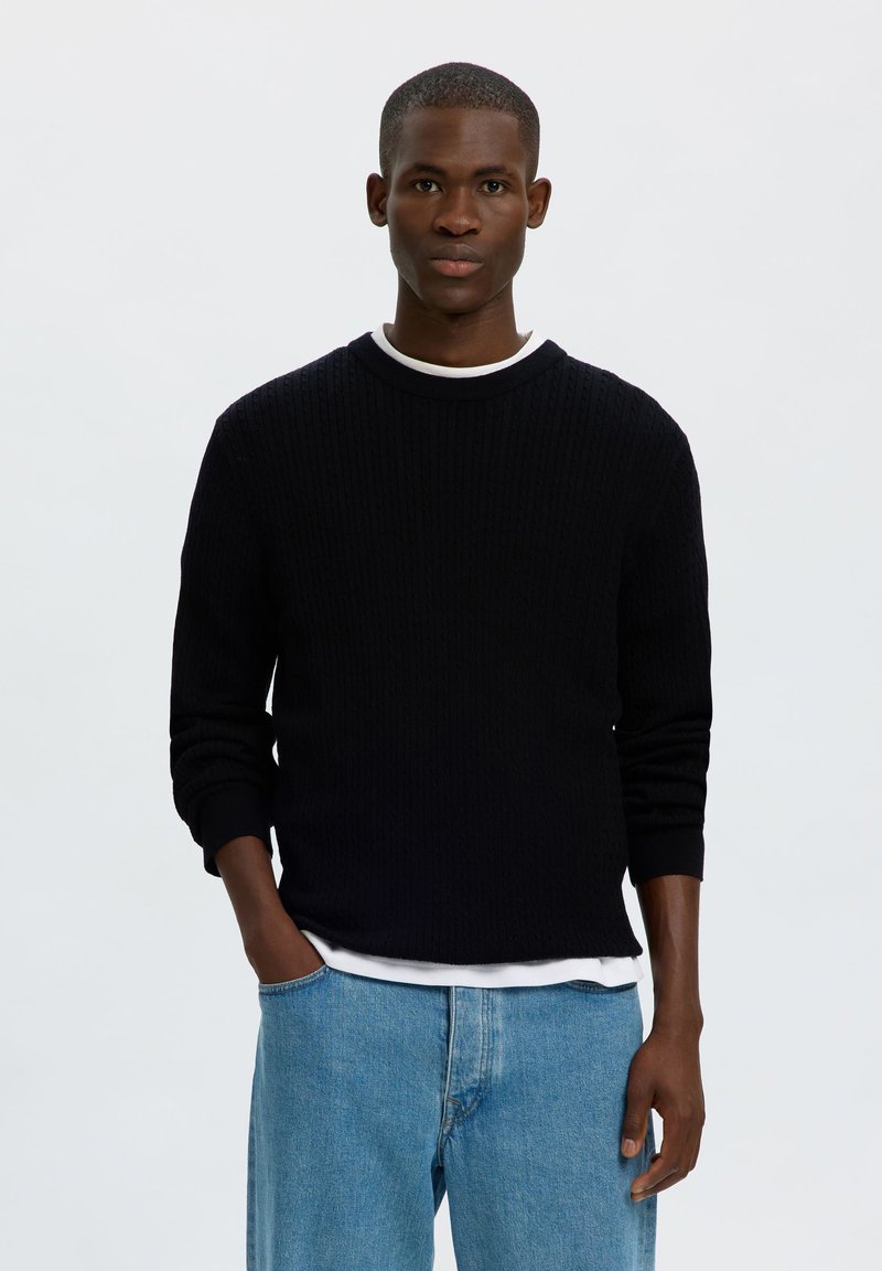 Jeune homme debout, mains dans les poches, portant un pull noir, un sous-vêtement blanc et un jean bleu clair sur un fond clair uni.
