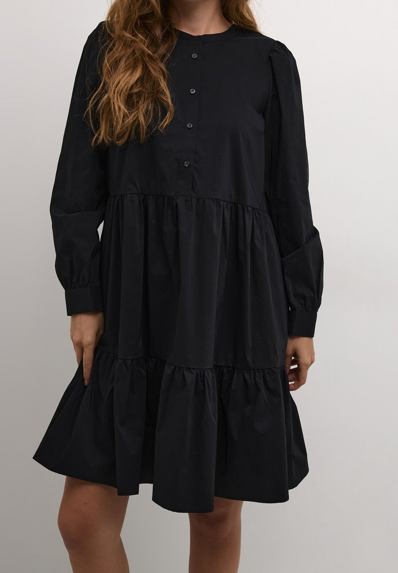 Femme portant une robe noire longue à volants avec des boutons sur la moitié supérieure, debout devant un fond clair uni.