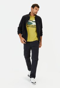 Marine zip-up jack met een groene T-shirt met een buitenthema tekst en zwarte cargo broek; witte sneakers maken de outfit compleet.